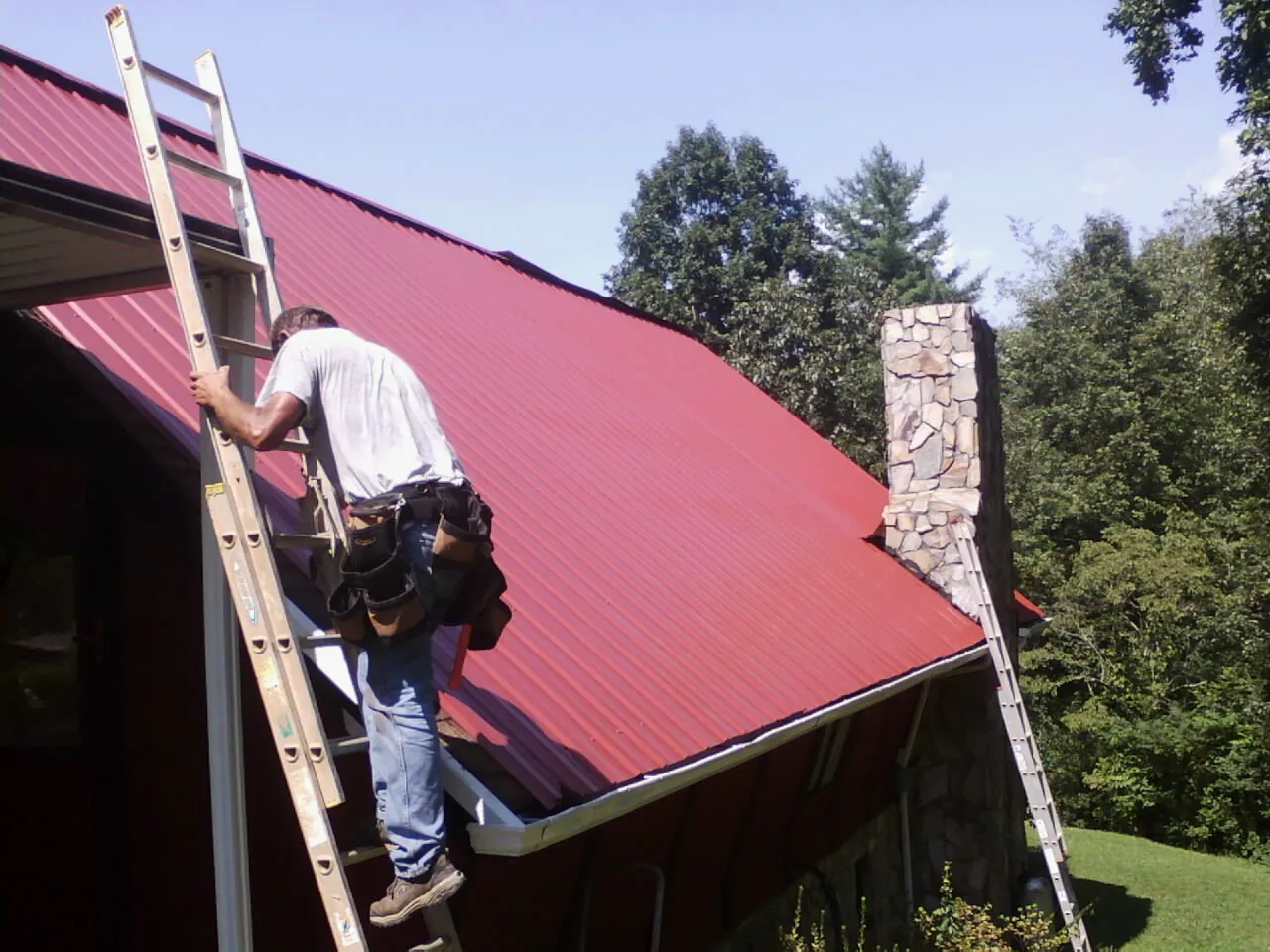 Roofer on a ladder beside a red metal roof and stone chimney — Soffit & Fascia Repair by Roofing Brothers in Coalton